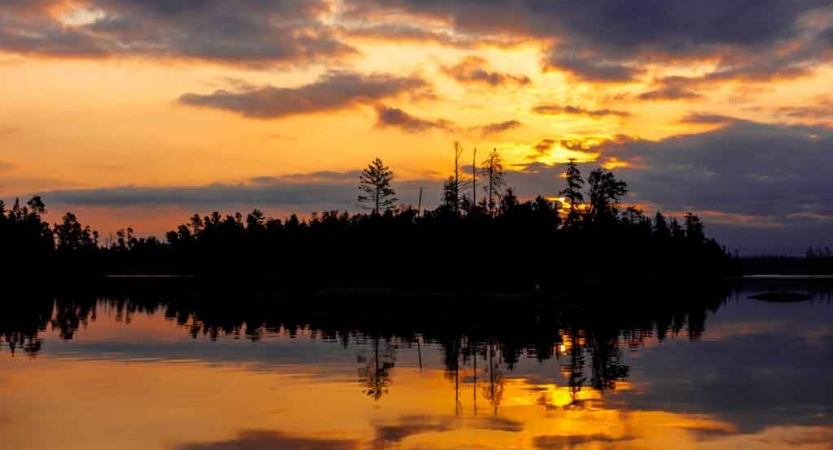 the sunset is reflected on a calm lake alongside a tree-lined shore